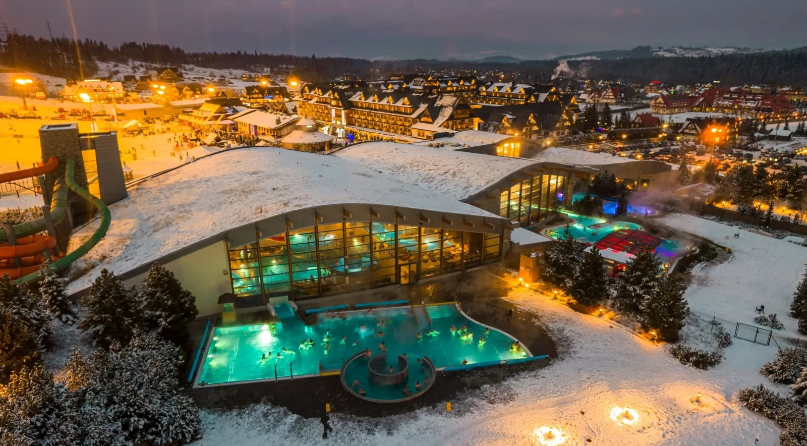 Lit outdoor thermal pools and a slide tower at dusk, with snow around the complex