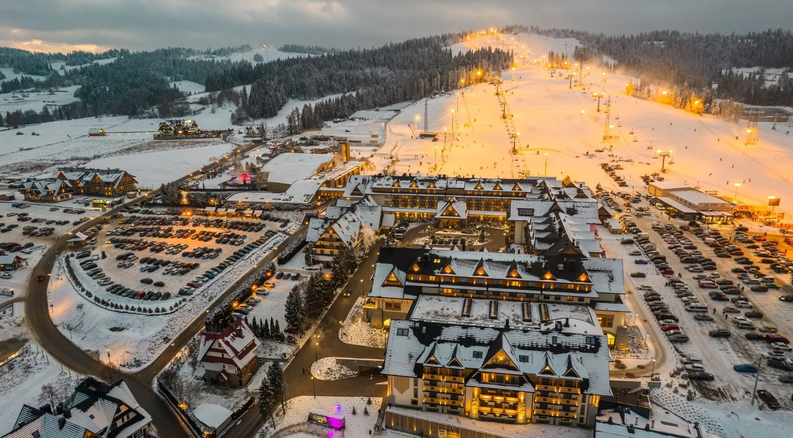 Aerial dusk view of a snowy mountain resort with a floodlit ski slope