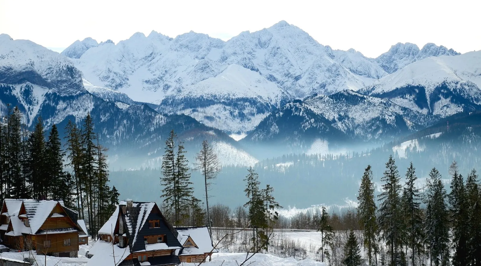 Snow-covered wooden highland chalets below a white mountain range in low mist