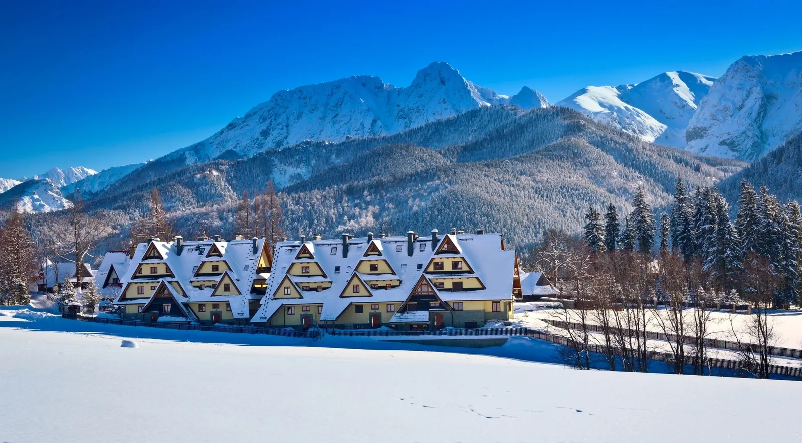 A row of snow-roofed highland-style buildings under bright winter peaks and a blue sky
