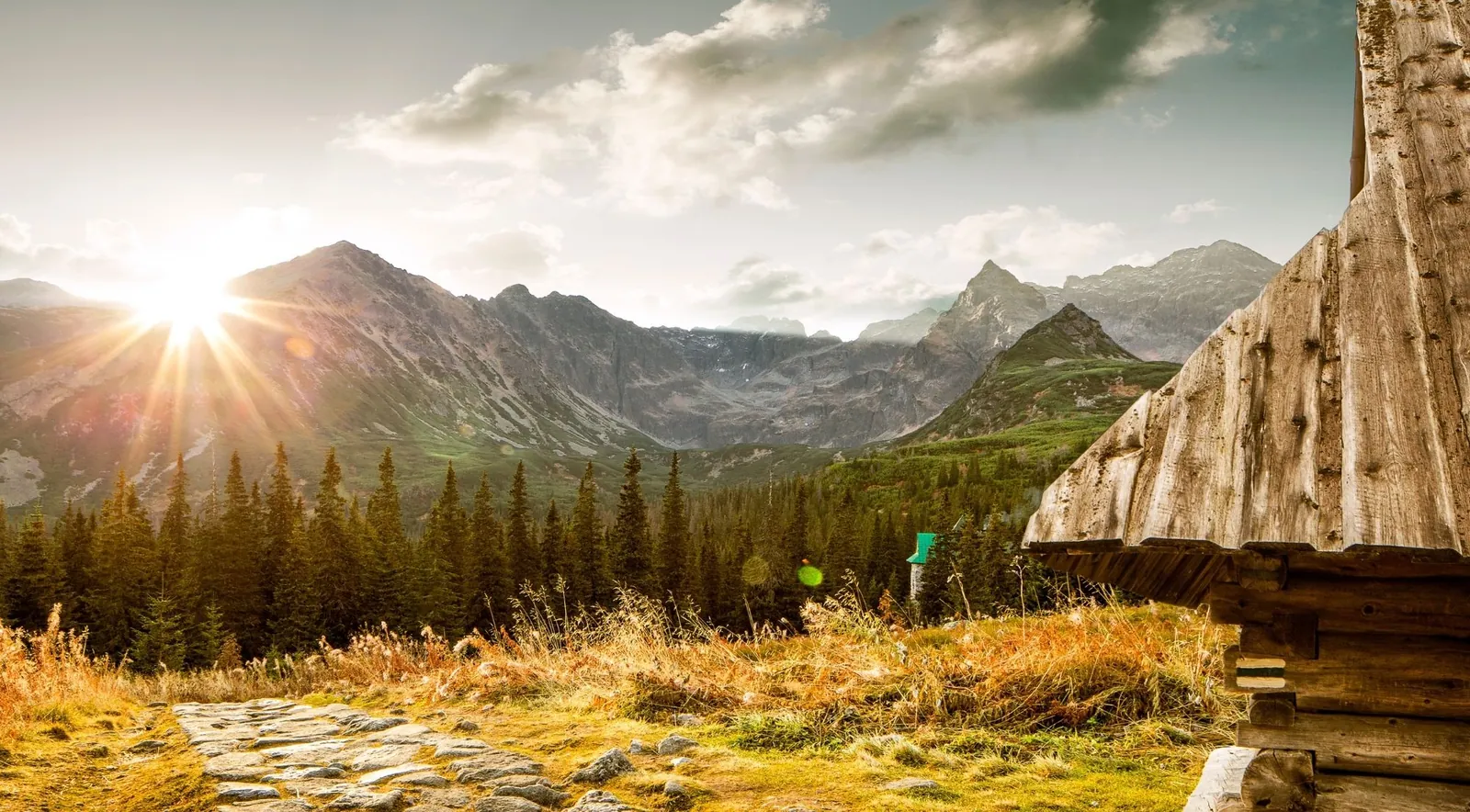 A mountain trail at sunrise beside a wooden shepherd's hut, with high peaks behind