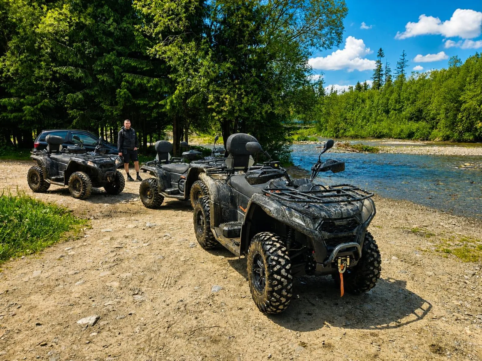 Three black quads parked on a gravel bank beside a clear river