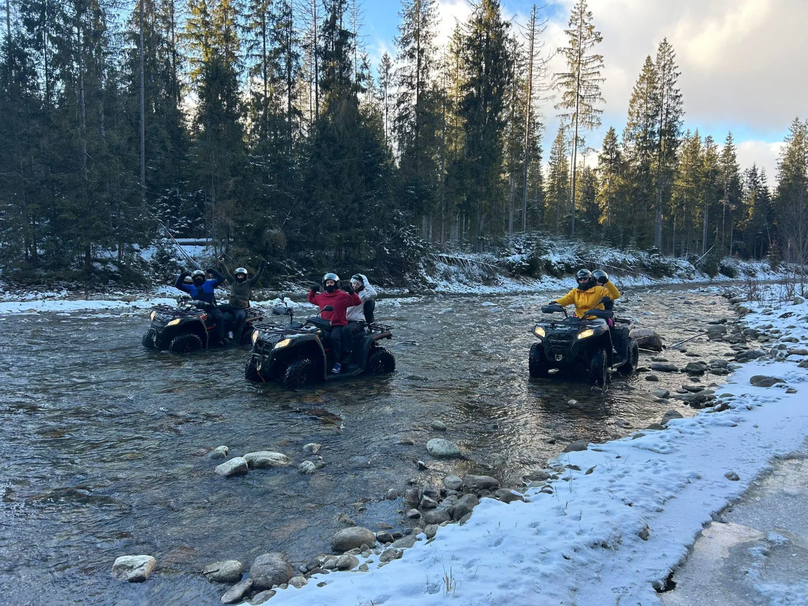 Riders on quads crossing a shallow river between snowy forest banks