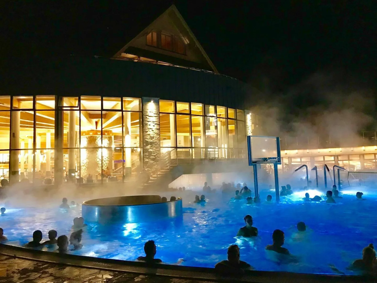 Bathers in a steaming outdoor pool at night beside a lit building