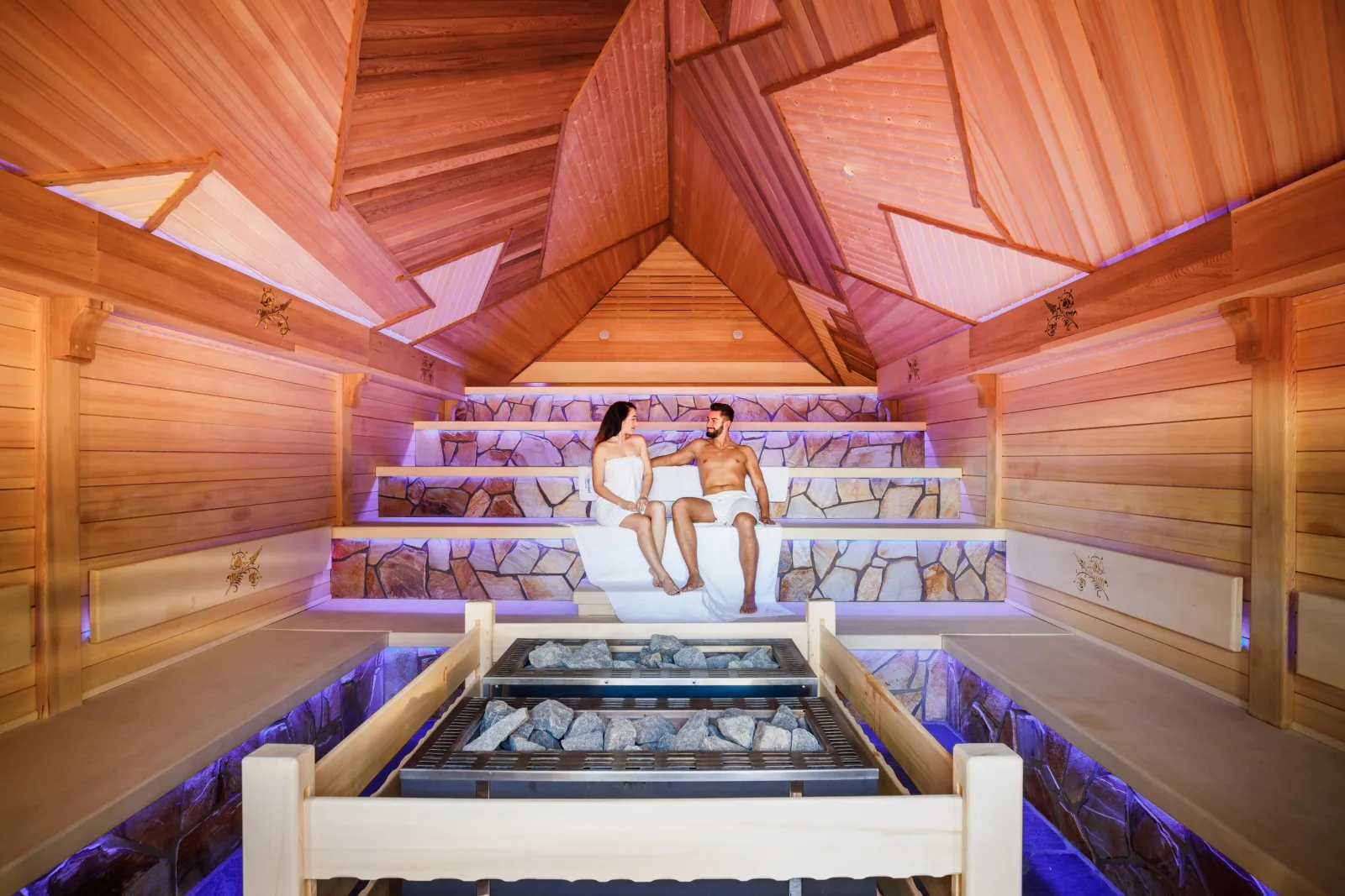 Couple in towels sitting in a large wooden sauna above stone heaters