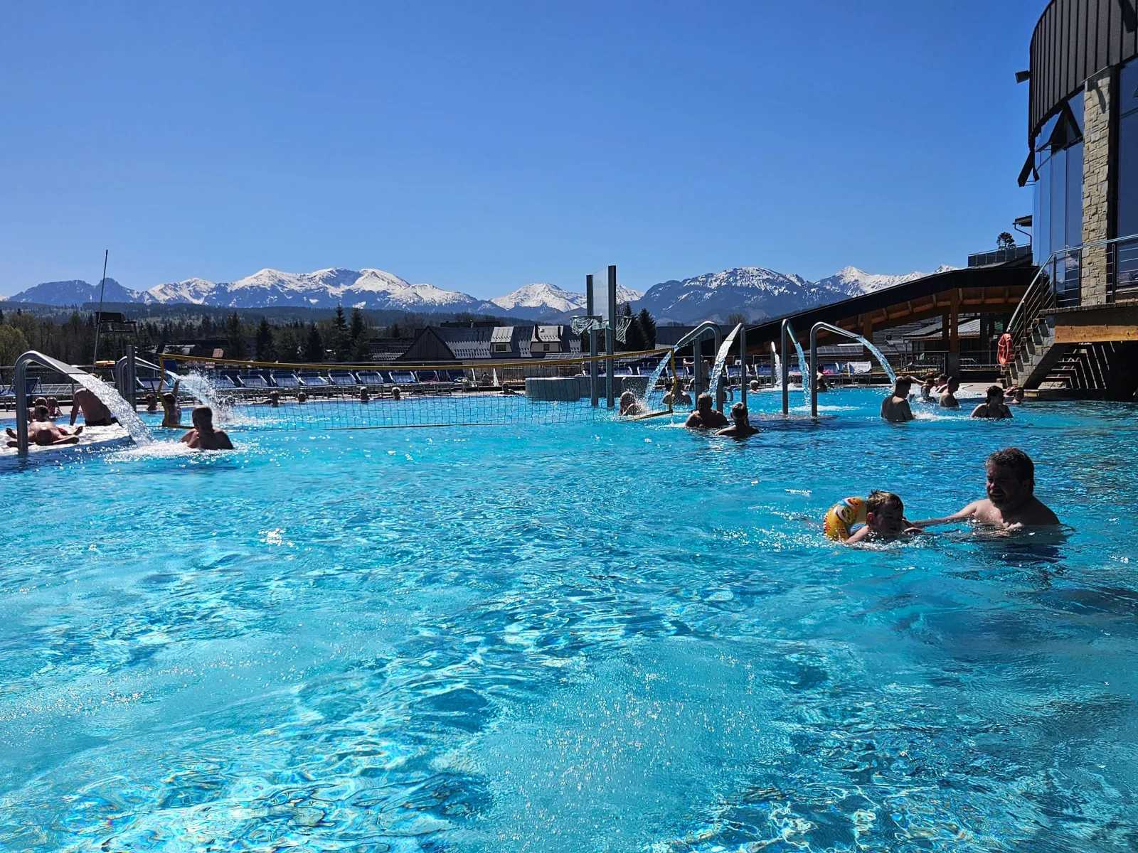 Swimmers in a sunlit outdoor pool with snow-capped mountains behind