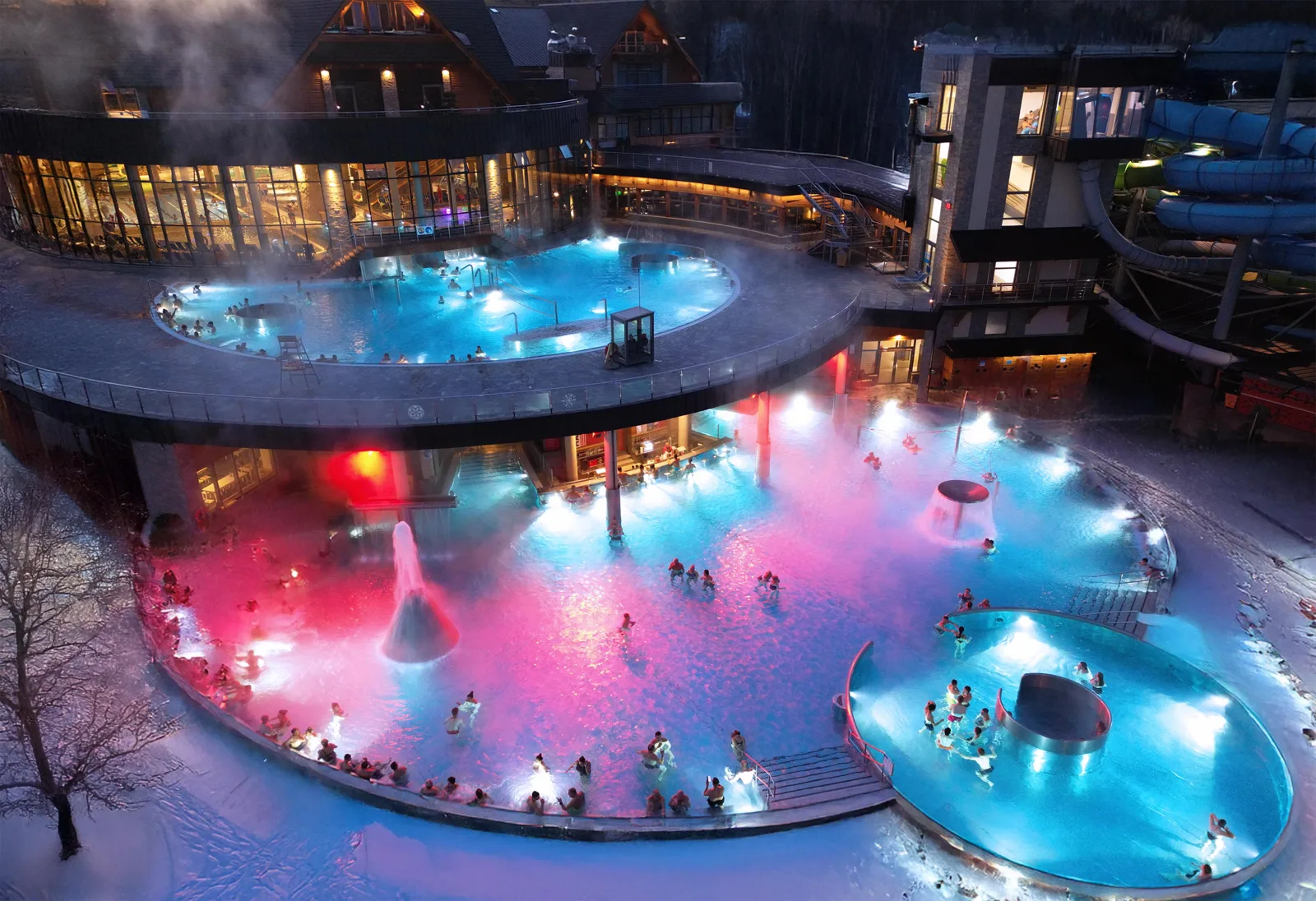 Steaming outdoor thermal pools lit pink and blue in the snow at night