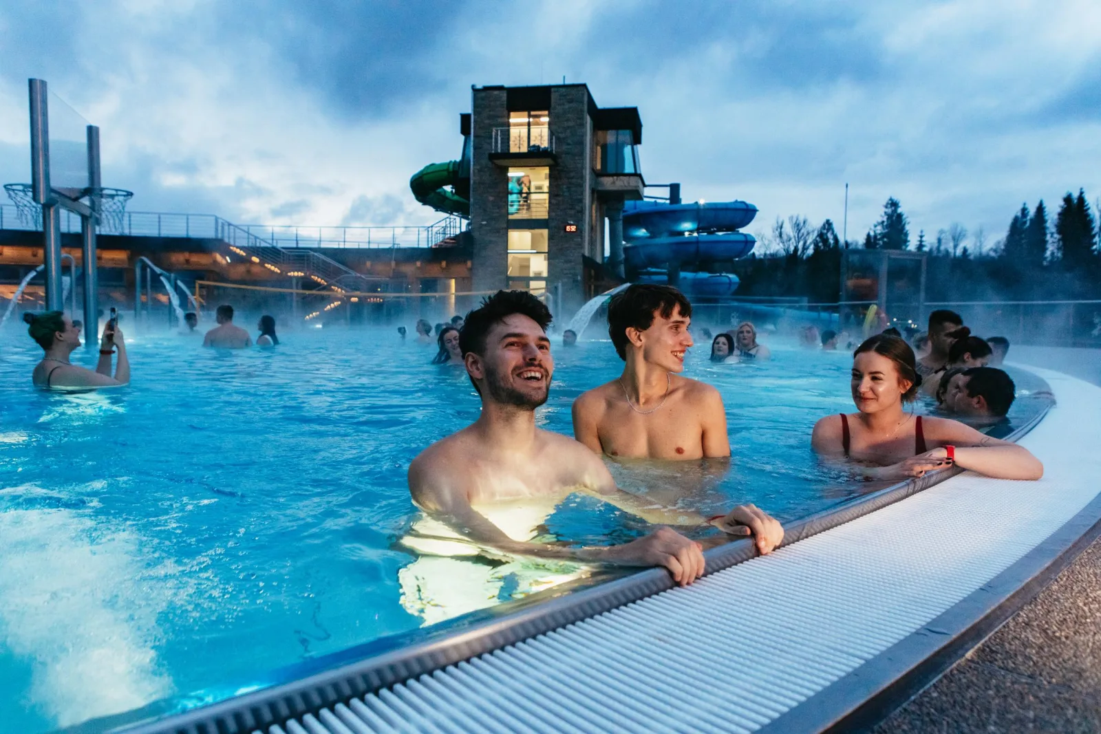 Friends relaxing in a steaming outdoor pool with water slides behind