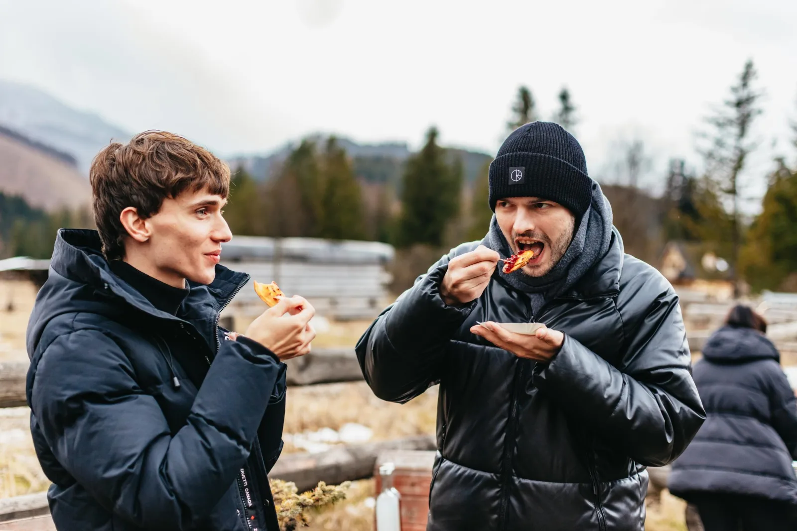 Two travellers eating grilled cheese outdoors near a mountain meadow