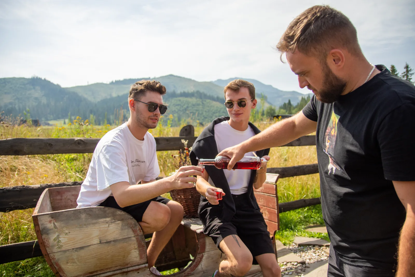 Host pouring red liqueur for two guests seated on a wooden bench