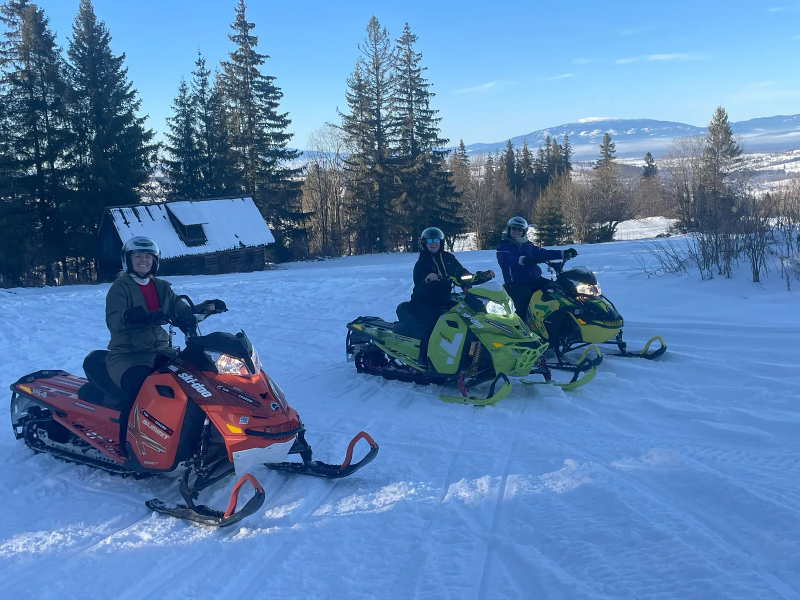 Three smiling riders on snowmobiles beside a snow-roofed wooden hut