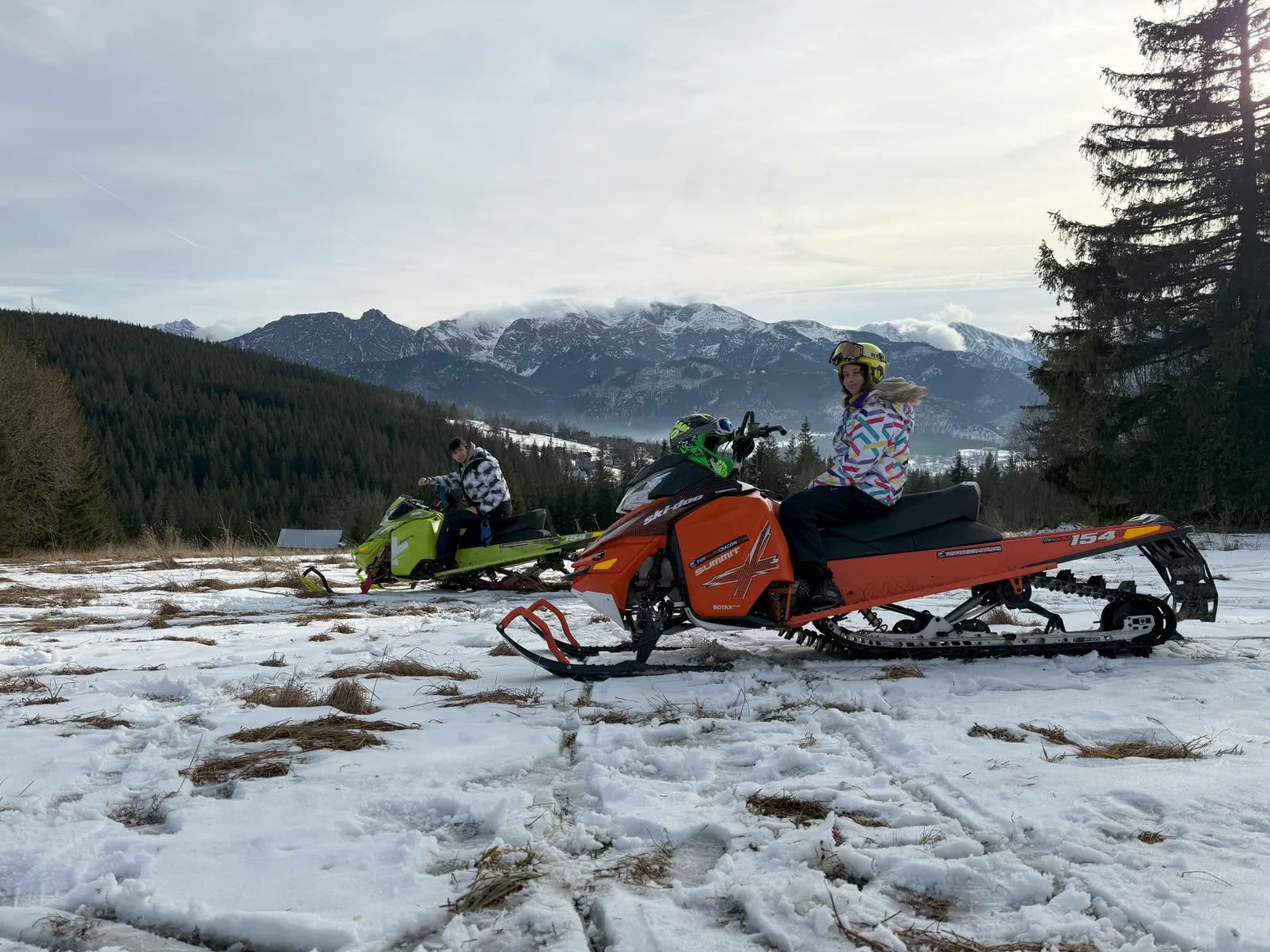 Two snowmobiles on a patchy snowy meadow below rocky mountains