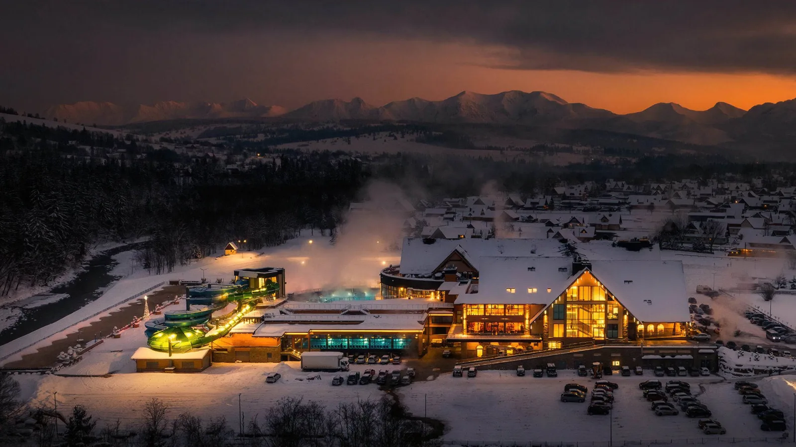 Snow-covered thermal baths complex lit up at dusk below mountains
