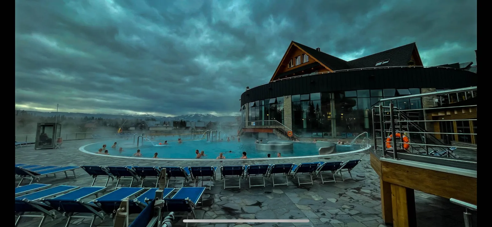 Steaming outdoor thermal pool with swimmers and empty sun loungers