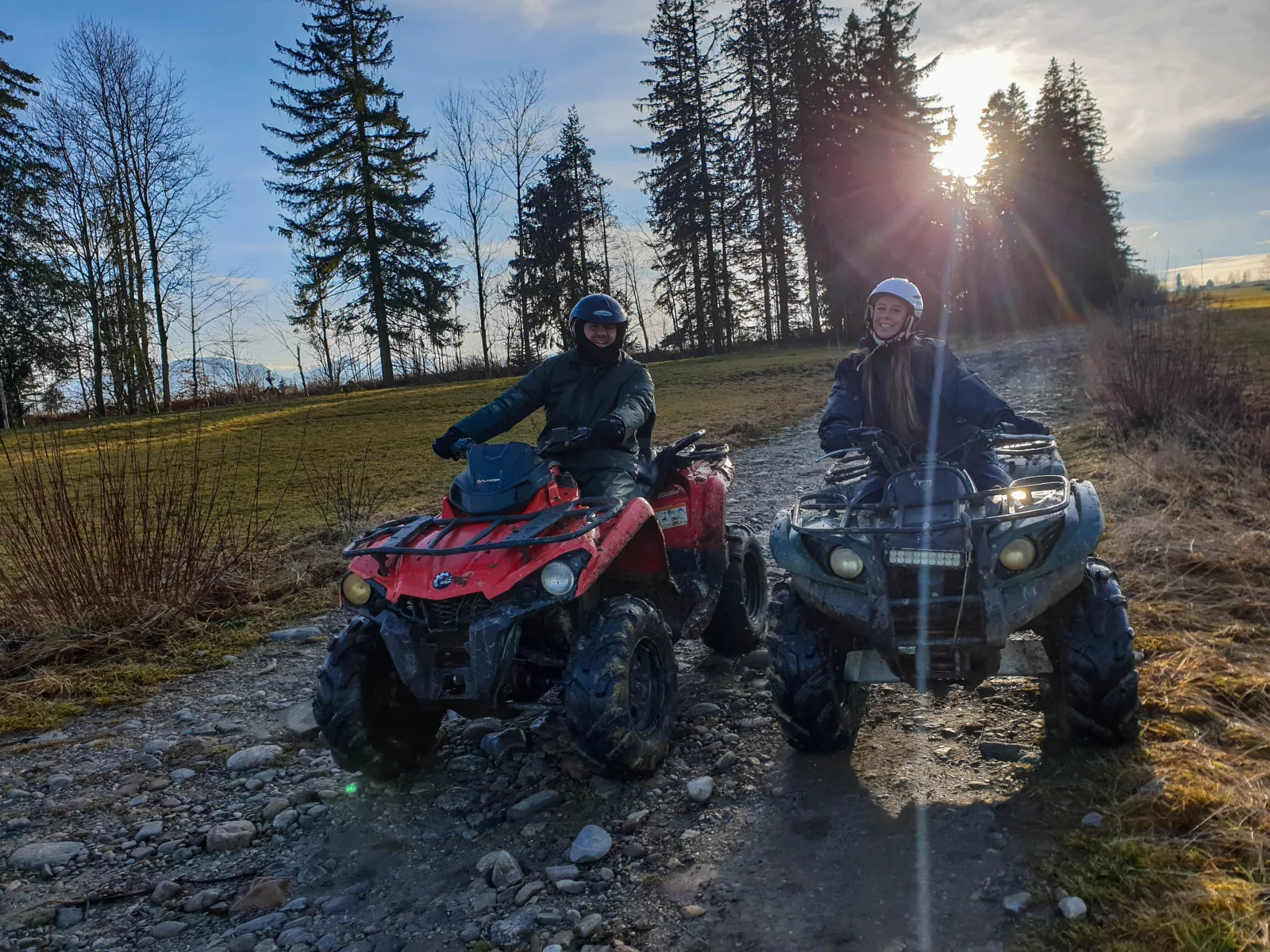 Two smiling riders on muddy quads on a stony track at sunset