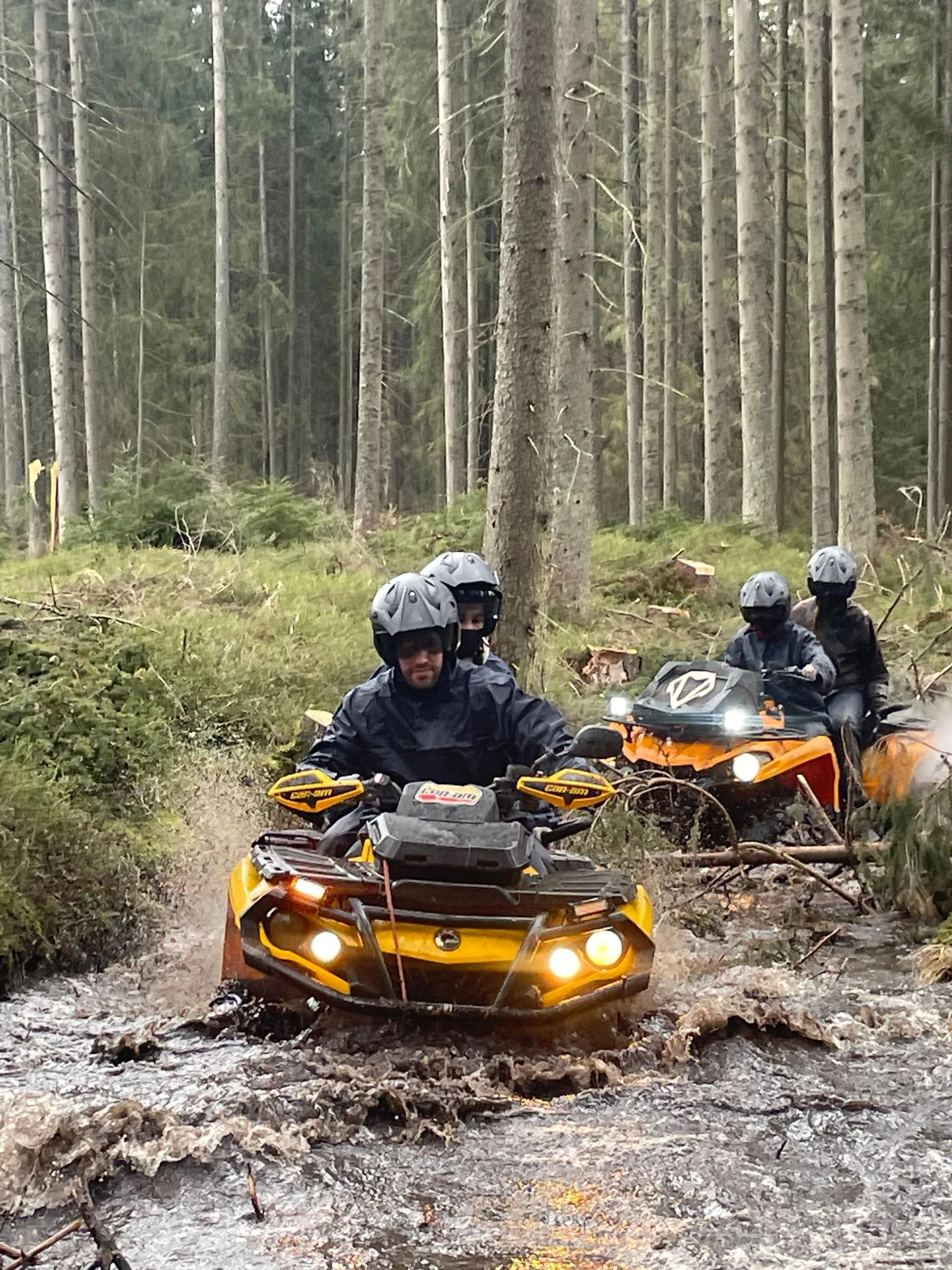 Helmeted riders on yellow quads splashing through a muddy forest stream