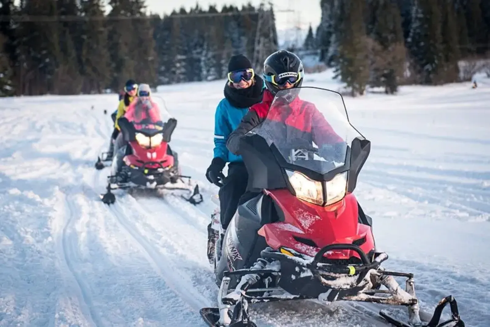 Riders in goggles driving red snowmobiles in a line across snow