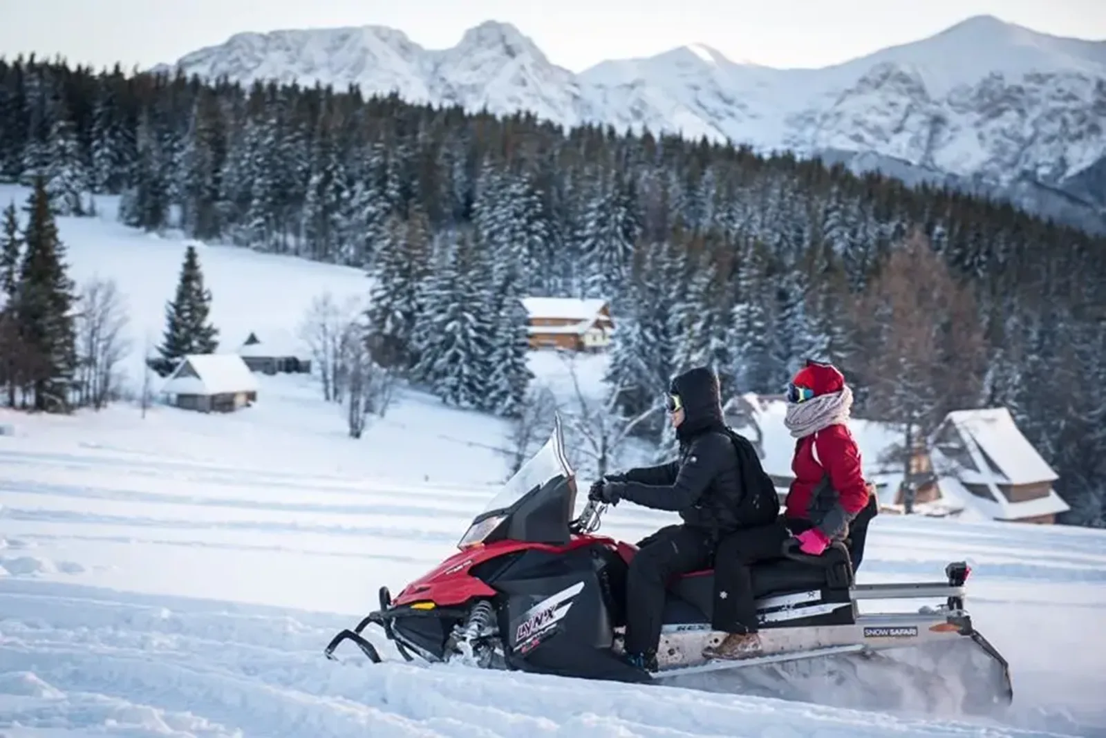 Two riders on a red snowmobile crossing a snowy meadow below mountains
