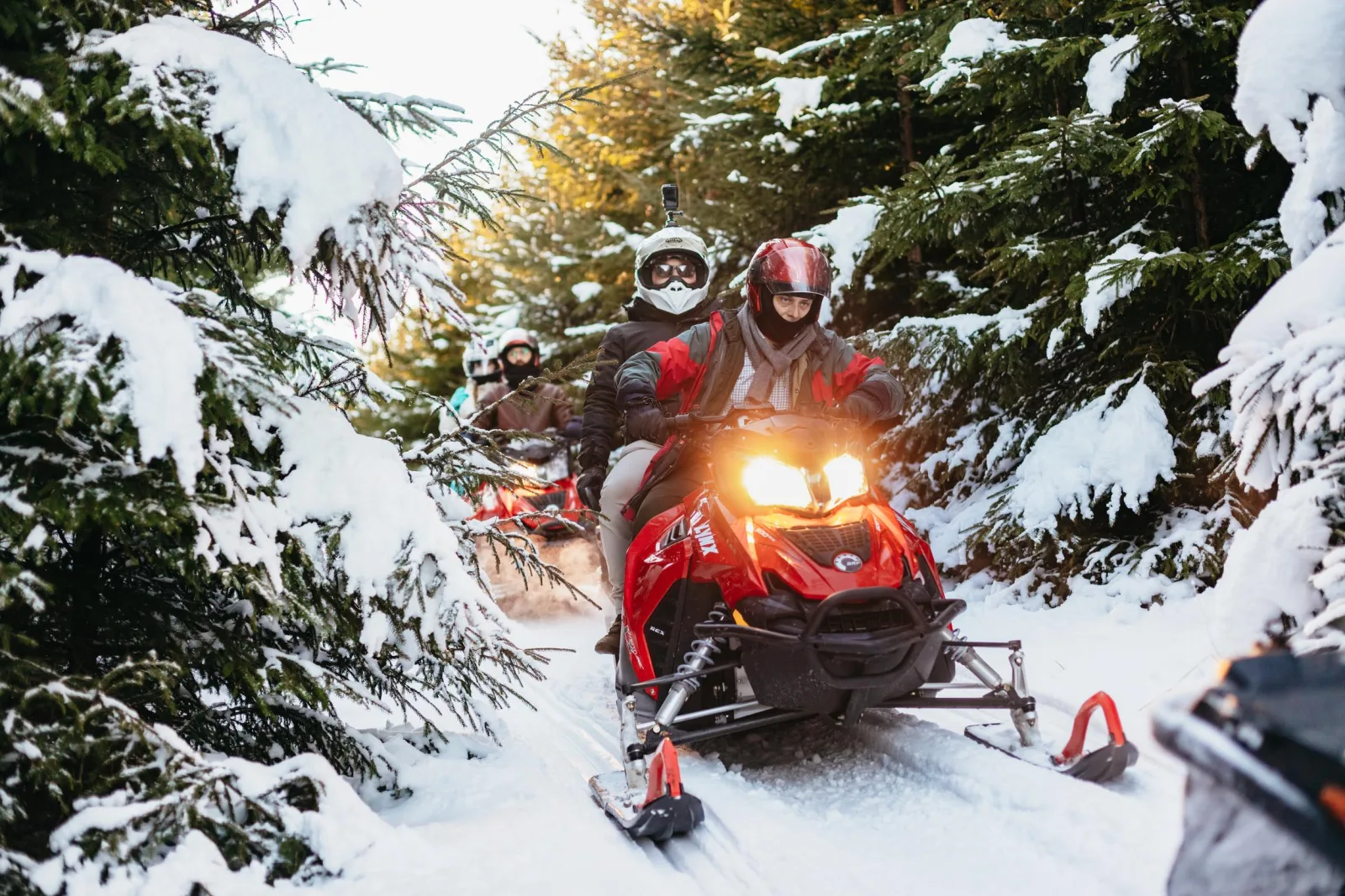 Snowmobile riders in helmets driving single file through a snowy forest