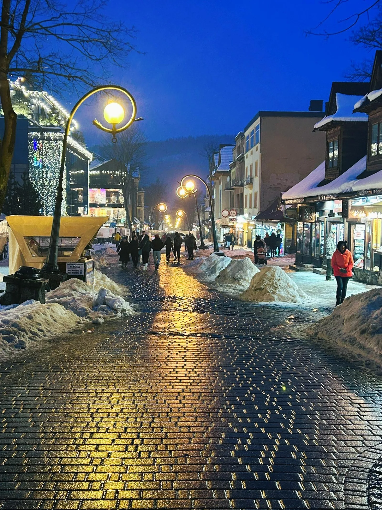 Lamp-lit pedestrian street with snow piles and evening shoppers