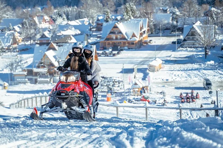 Two riders on a red snowmobile climbing a snowy slope above wooden chalets