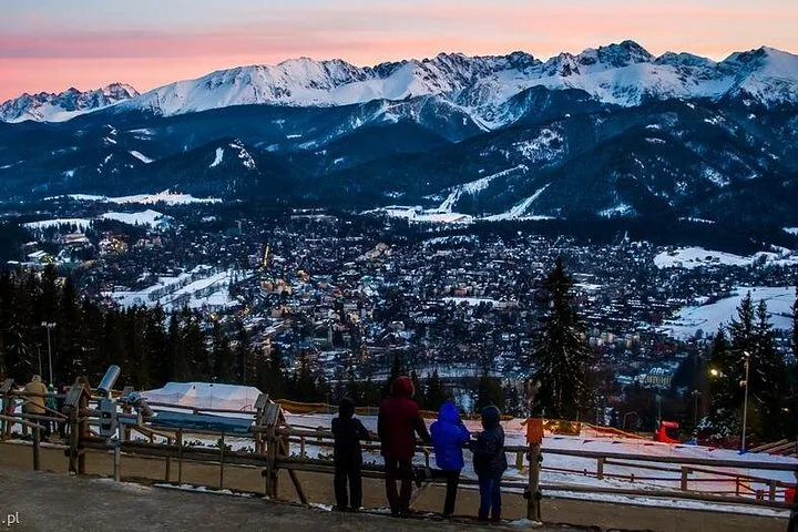 Visitors at a railing watching sunset over snowy Tatra peaks and a lit town