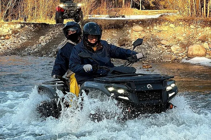 Two riders on an ATV splashing through a river in autumn light