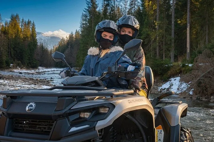 Two helmeted riders on a grey ATV beside a forest river